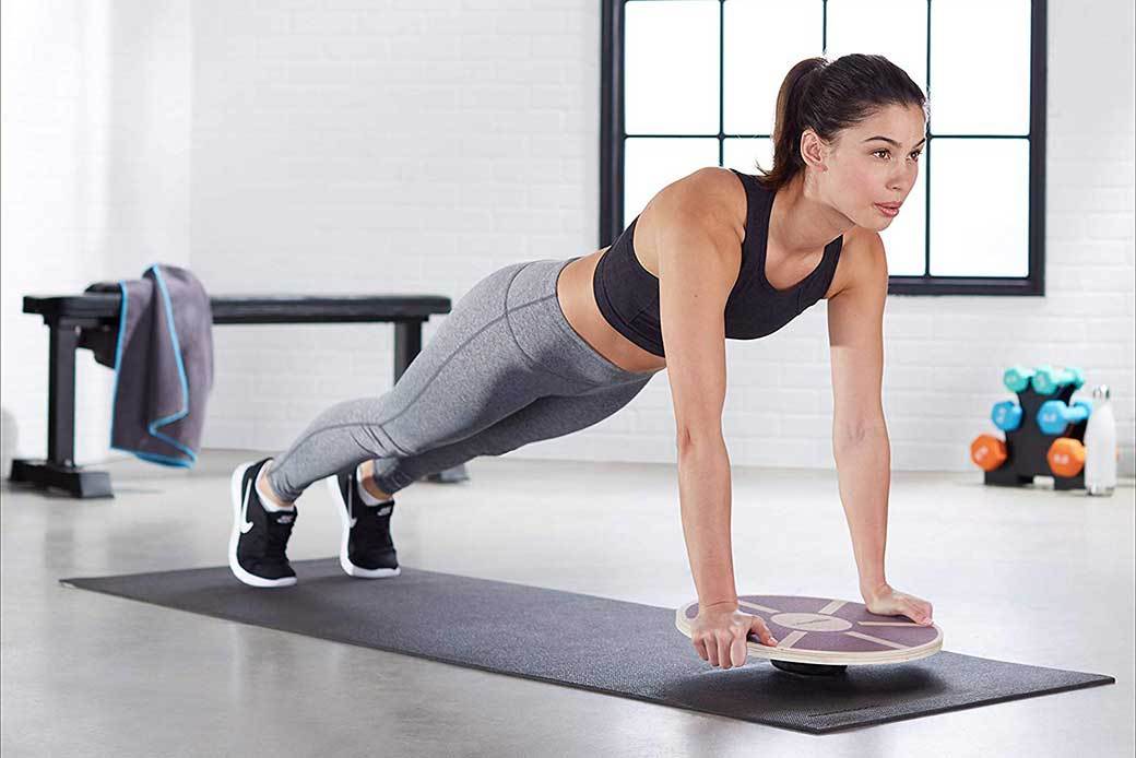 A woman wearing workout gear is in a gray studio and using a wobble board during a push up.
